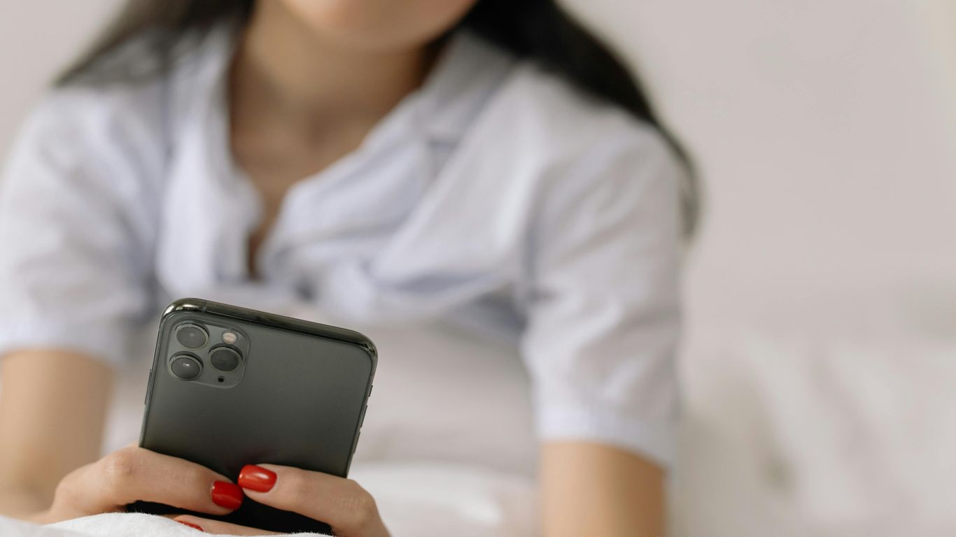 Person lying on bed, holding smartphone with red painted nails.