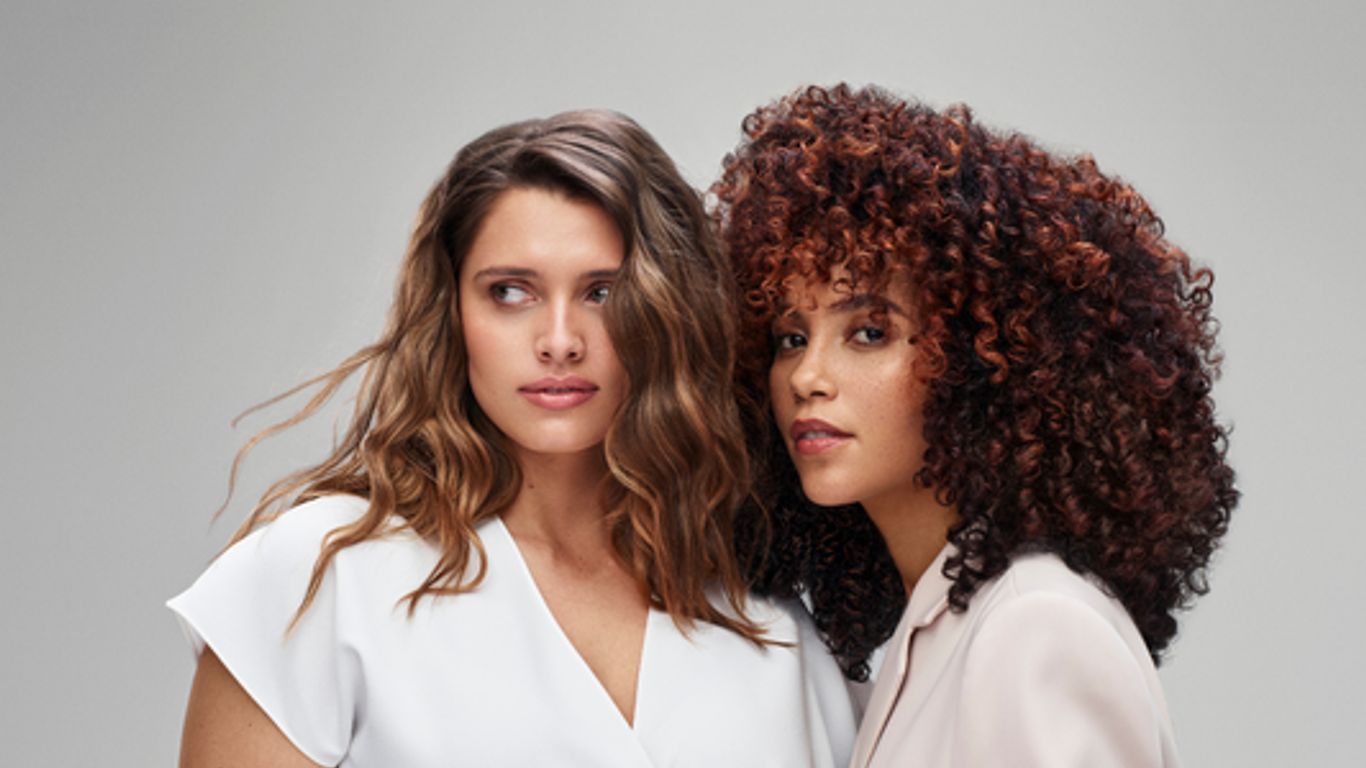 Two women posing confidently, one with curly hair, studio background.