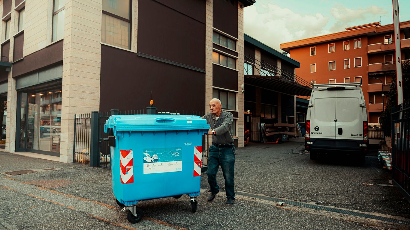 A man pushing a blue trash can down a street