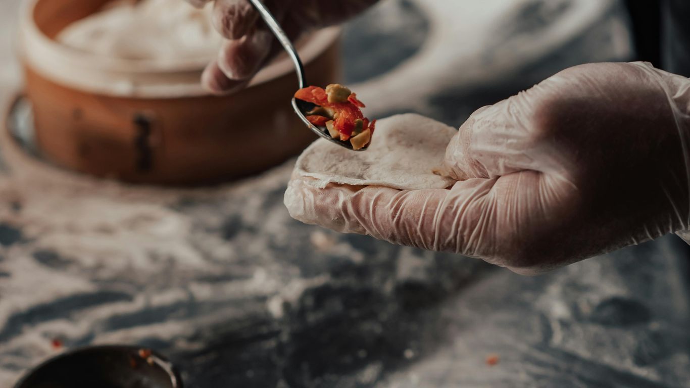 Gloved hands preparing dumplings with filling on a floured surface.