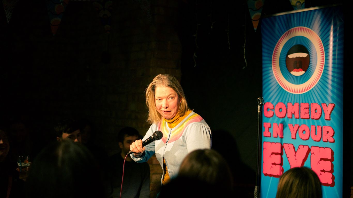 A comedian performs on stage with a "Comedy In Your Eye" banner.