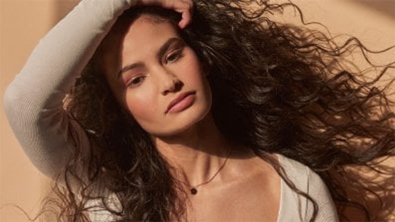 Person posing, long curly hair, subtle light, neutral colors.