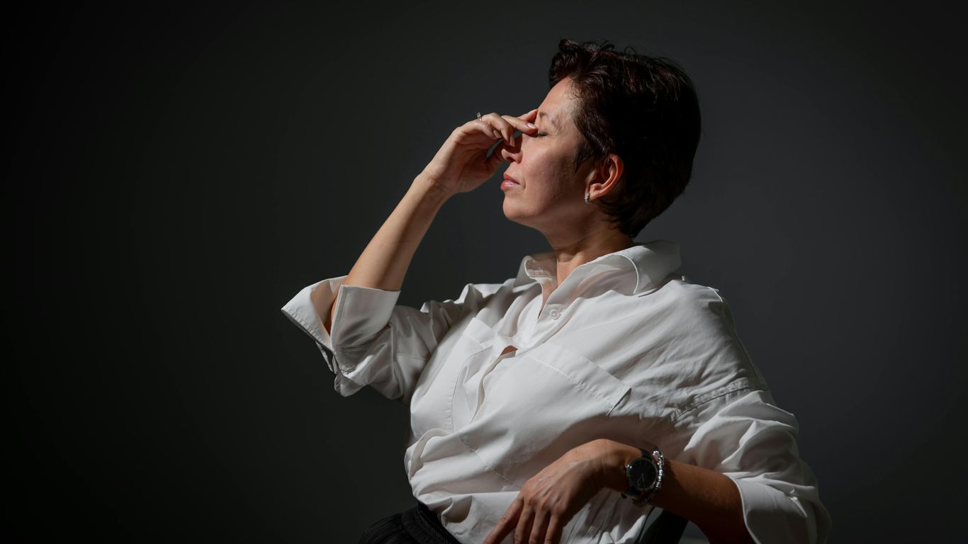 Person in white shirt sitting, pinching nose, looking stressed.