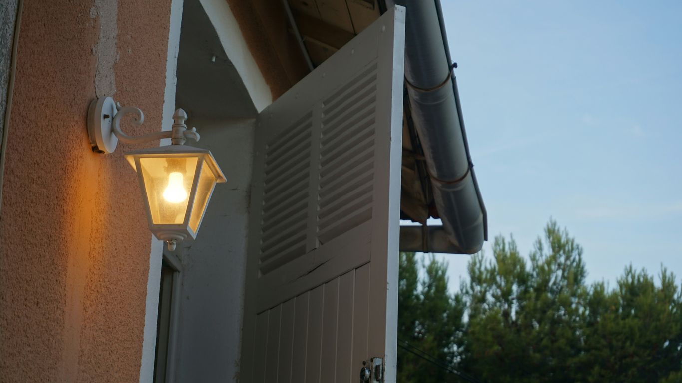 Outdoor lamp illuminates open white shutters on building.