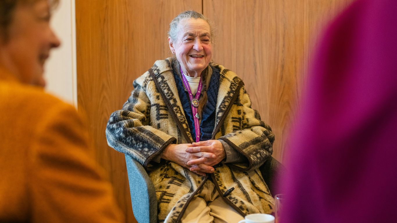 Elderly woman in patterned coat smiling at gathering.