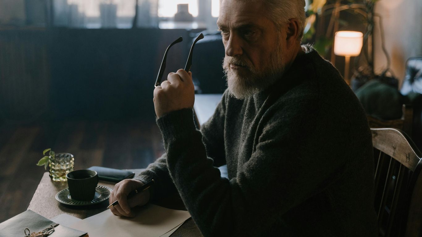 Person sitting at desk, holding glasses, thoughtful, cozy room.
