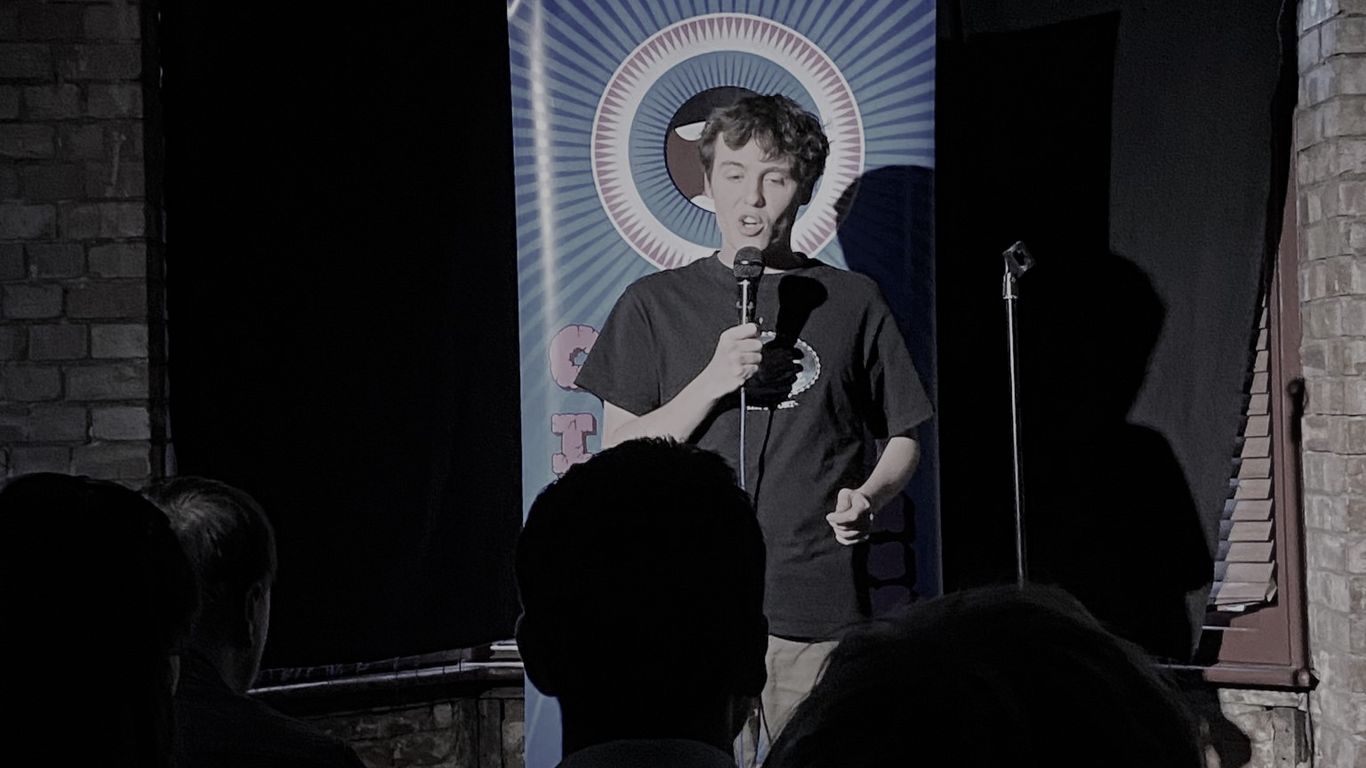 A person performs stand-up comedy on a stage with a banner.