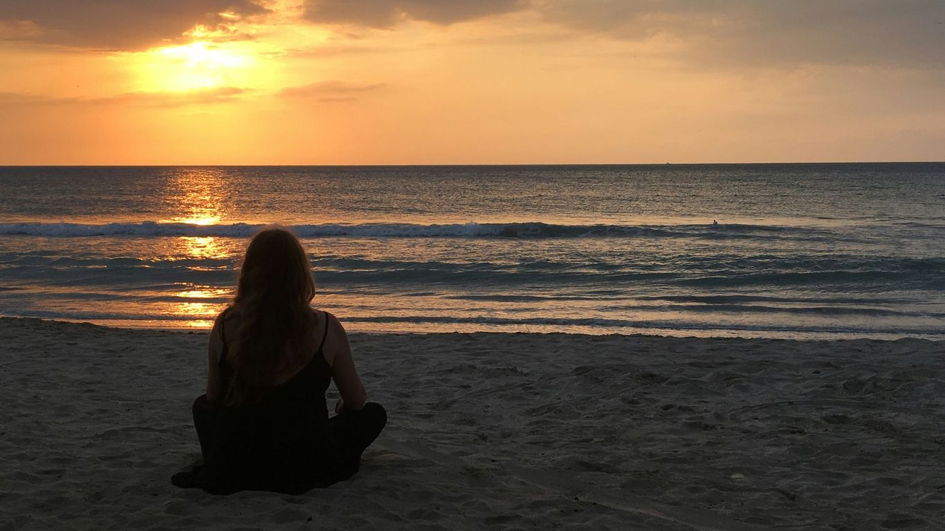 woman wearing black spaghetti strap top sitting and facing near seashore under orange sky