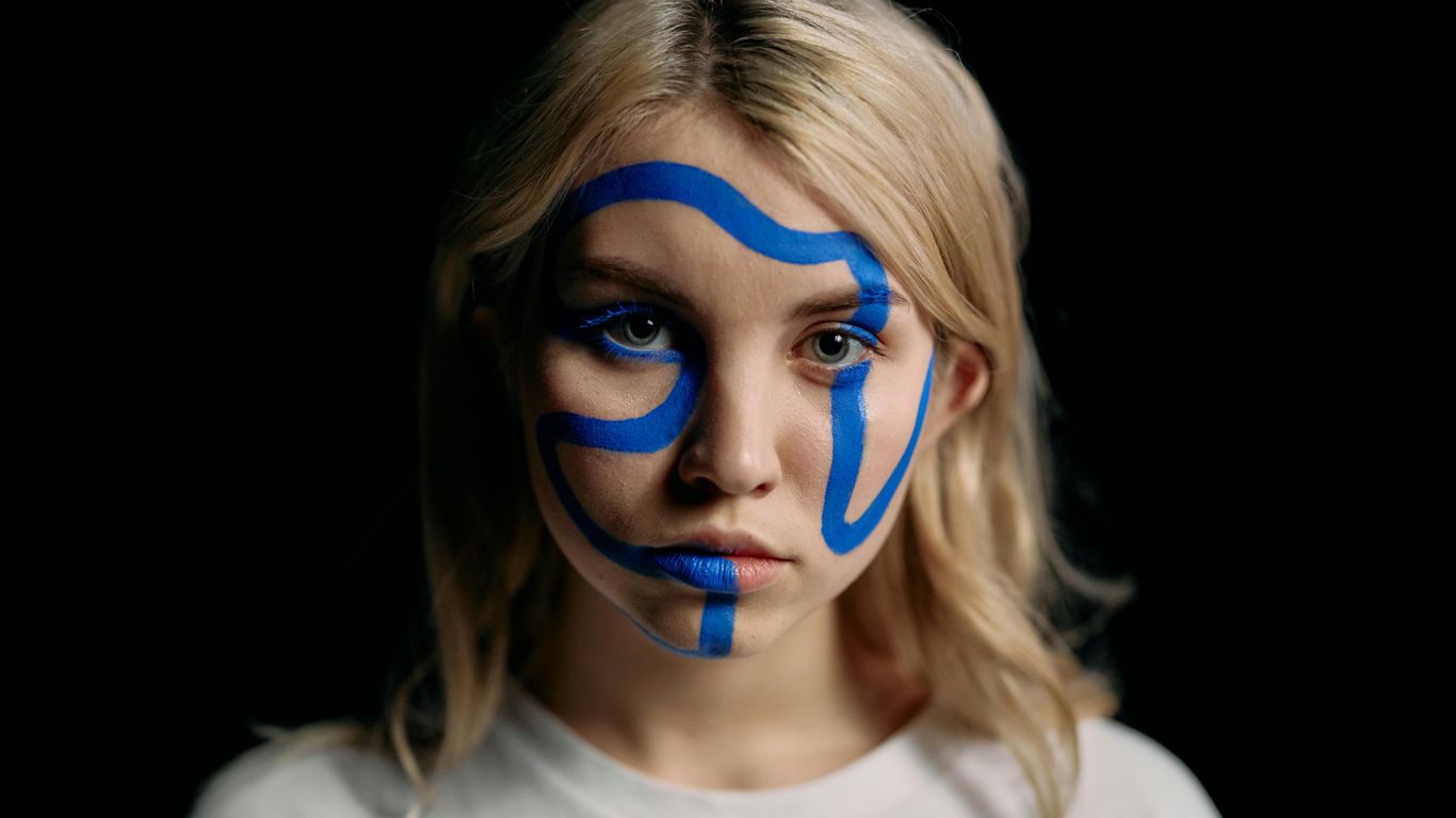 Person with abstract blue face paint, neutral expression, dark background.