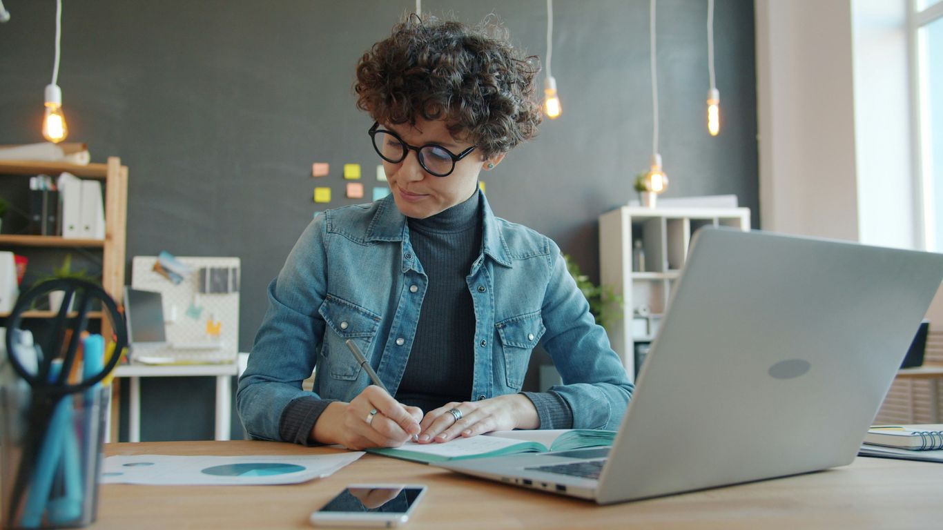 Woman with curly hair writing at desk with laptop