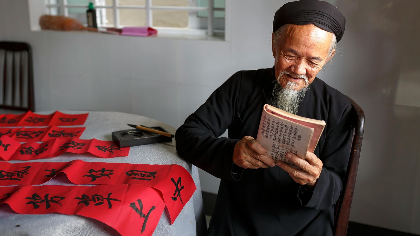 An elderly person reads a book next to red calligraphy scrolls.