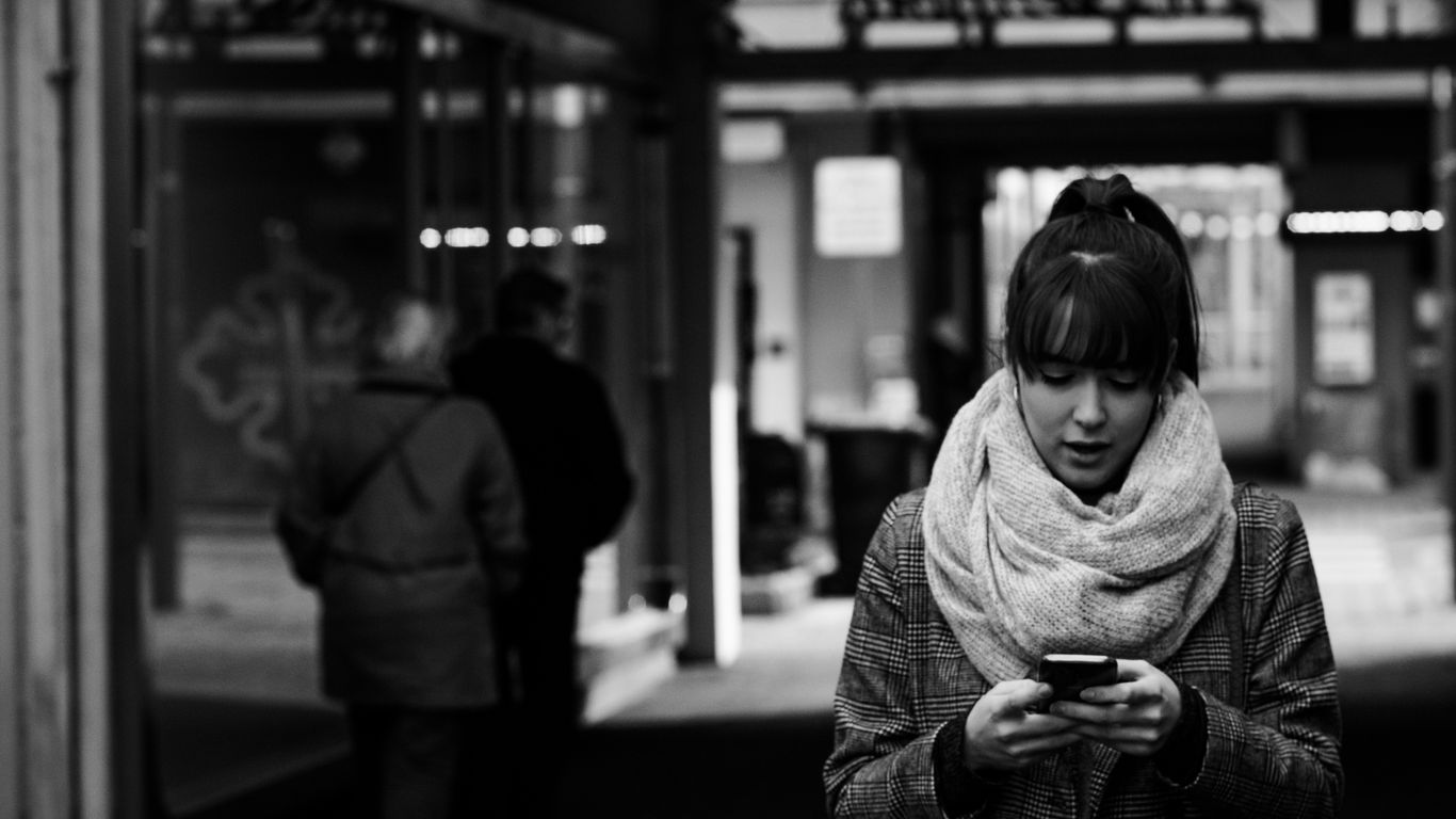 grayscale photography of woman walking near street while using smartphone