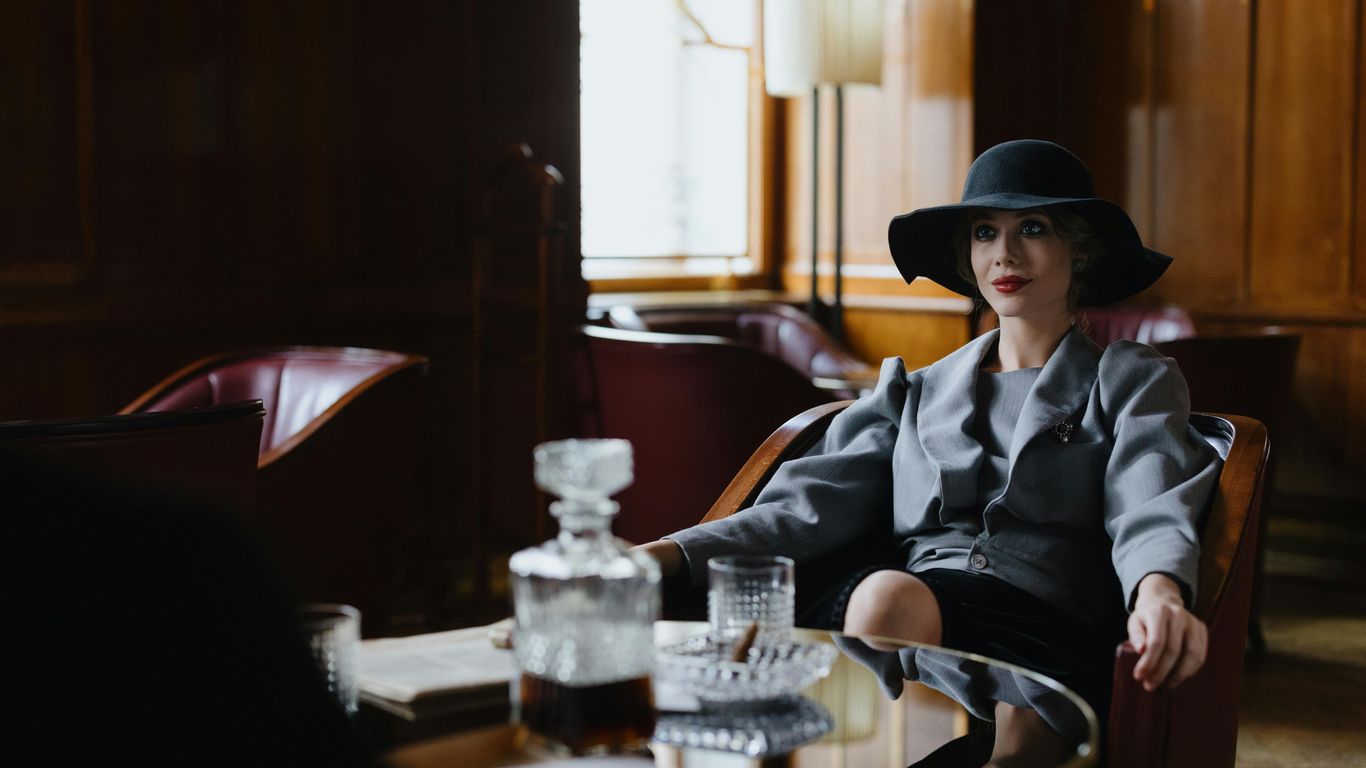 A person in a hat sits in a dimly lit room with a decanter.