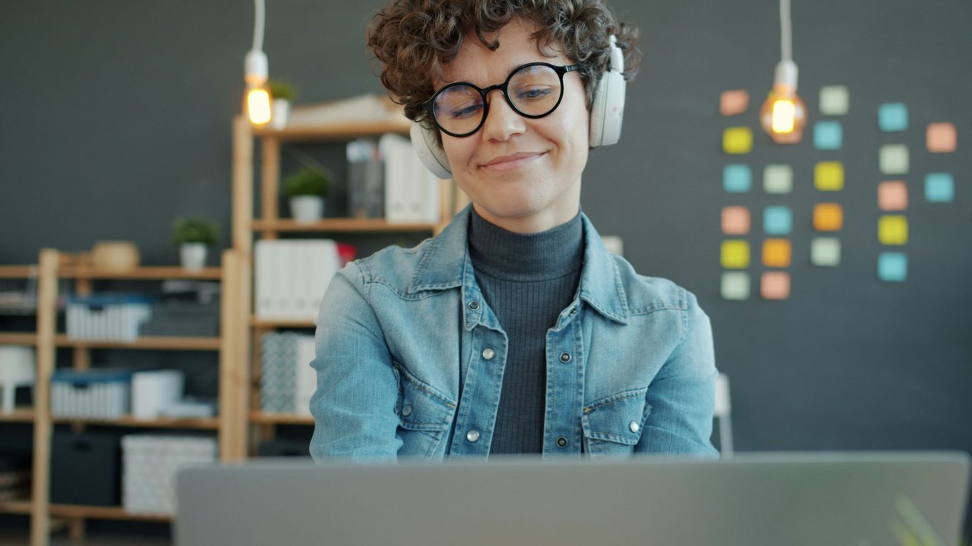 Woman wearing headphones smiles while looking at laptop.