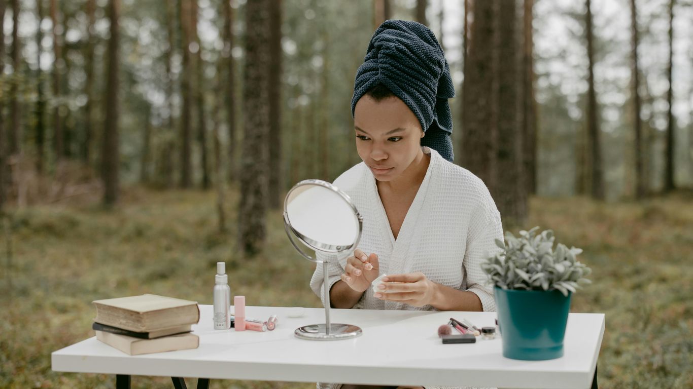 Person sitting at table in forest, applying skincare, mirror nearby.