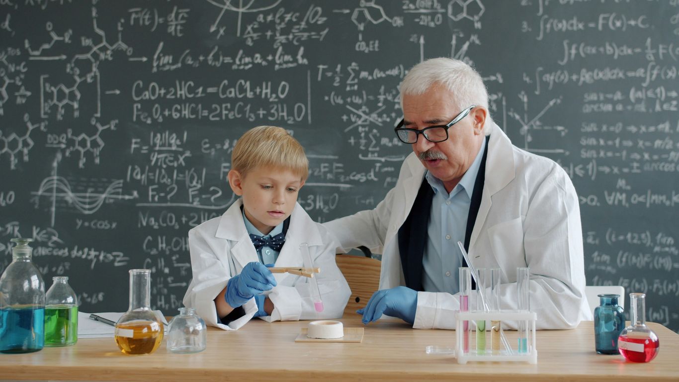 Elderly scientist teaches young boy in lab coat.