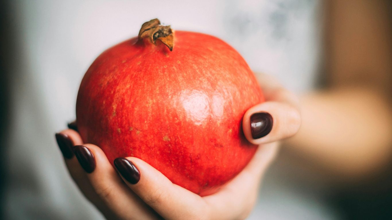 A hand holding a ripe pomegranate with dark nail polish.