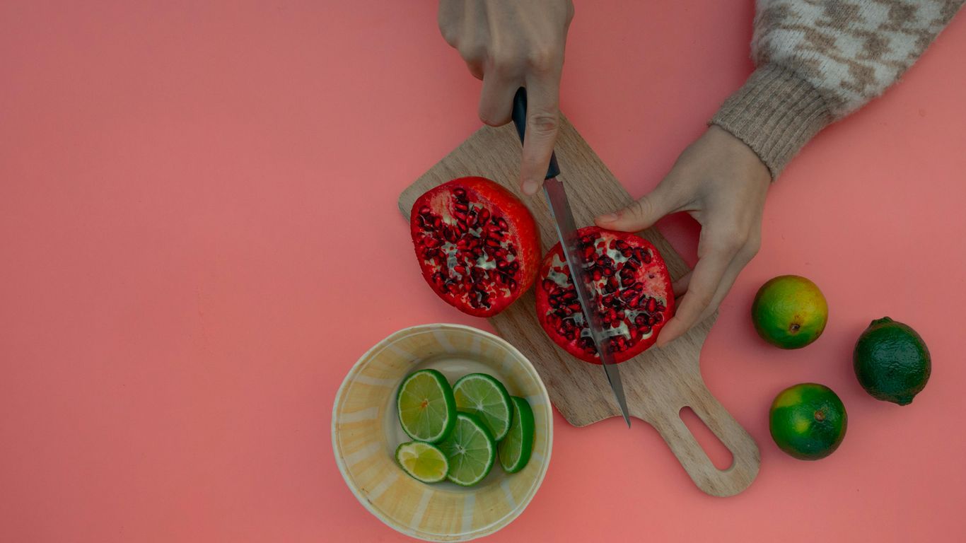 Hands cutting pomegranate, limes in bowl, pink background, wooden board.
