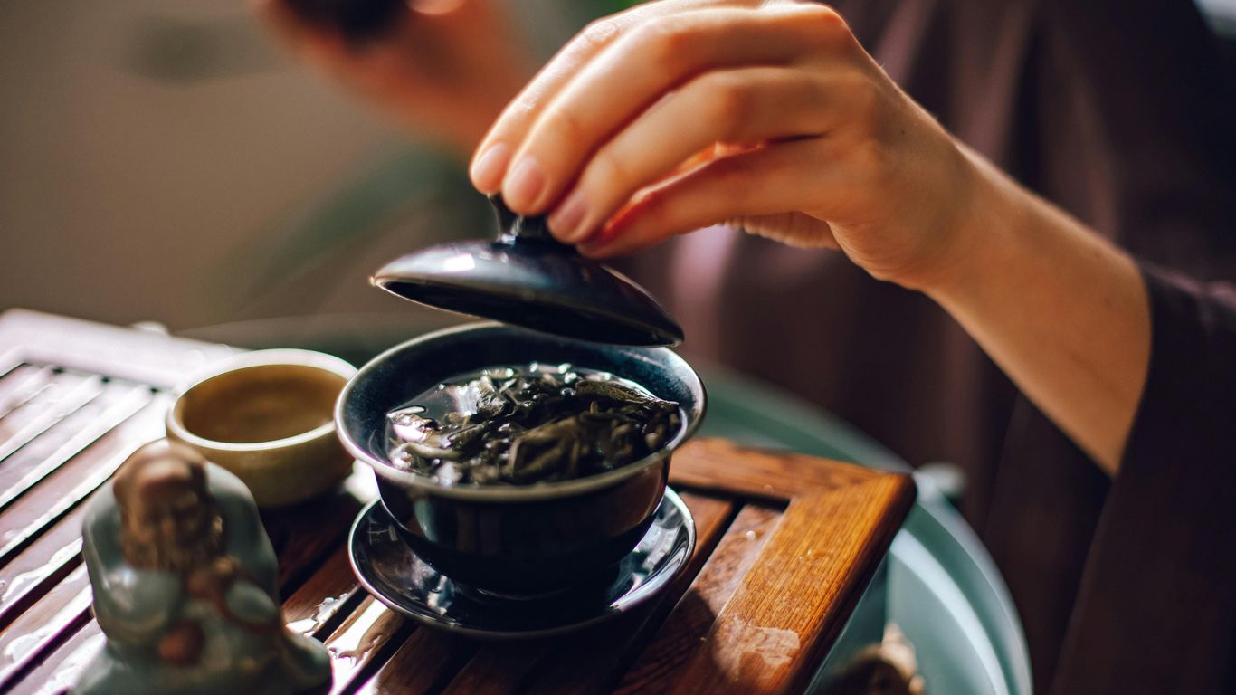 A person prepares tea with a small figurine on a wooden tray.