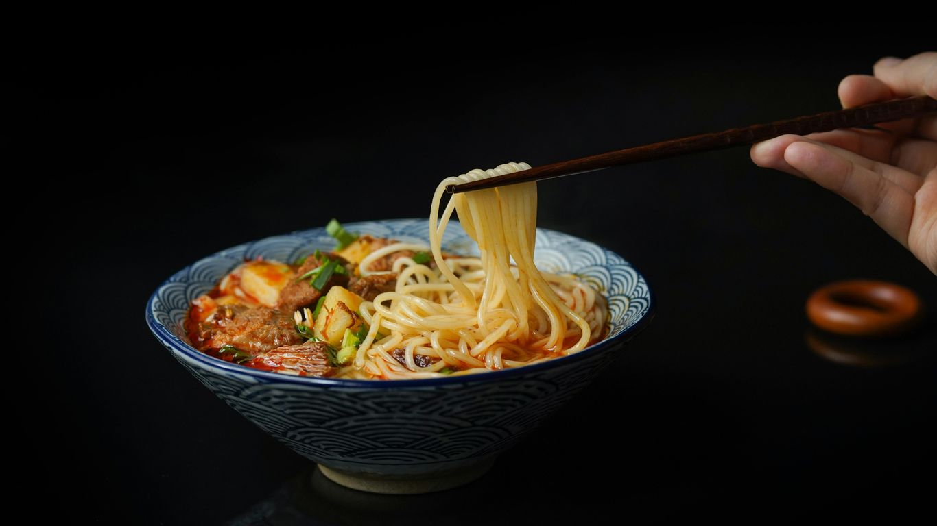 Bowl of noodle soup with chopsticks lifting noodles, dark background.