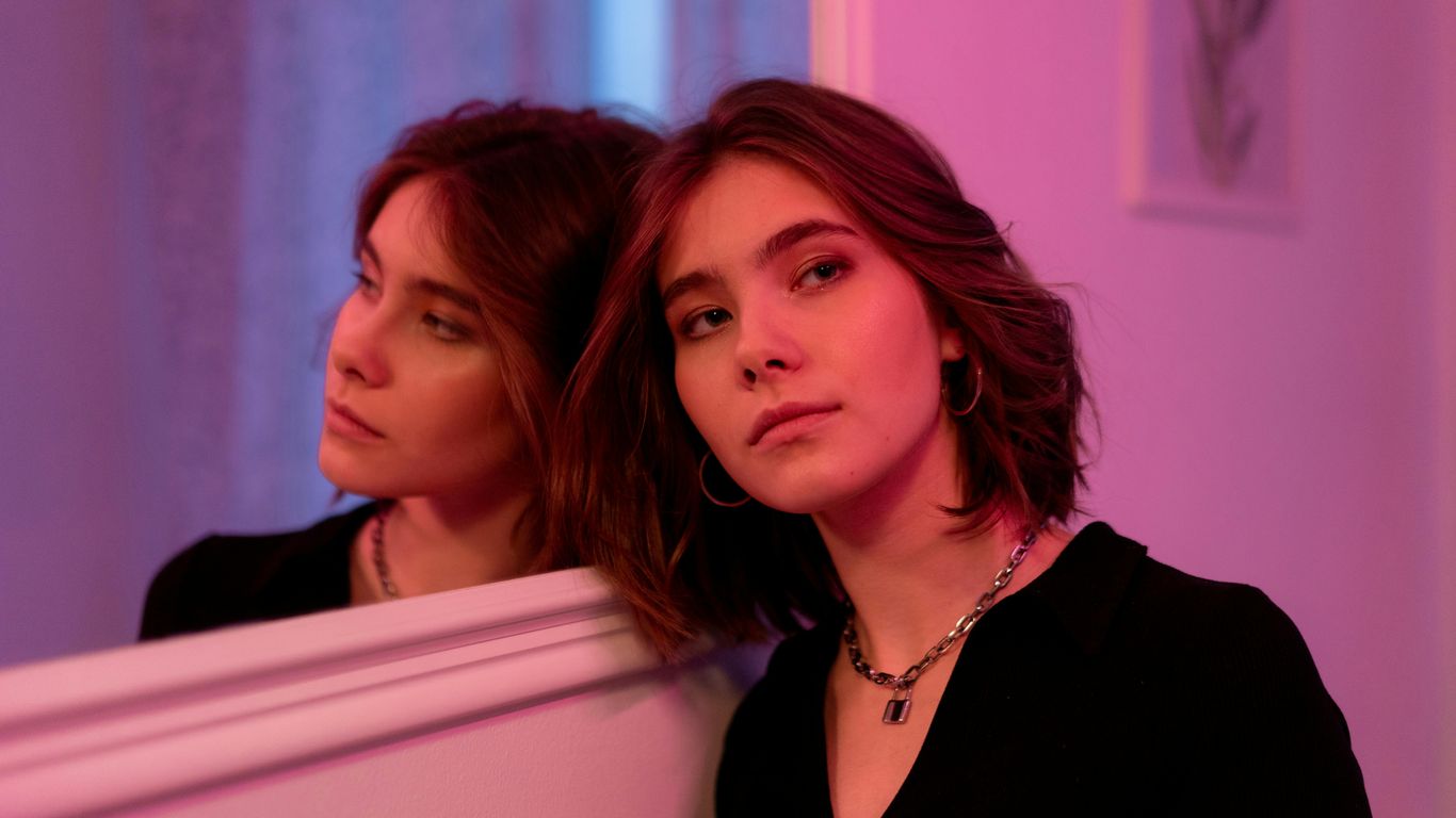 Person leaning against mirror, pink lighting, thoughtful expression, casual outfit.