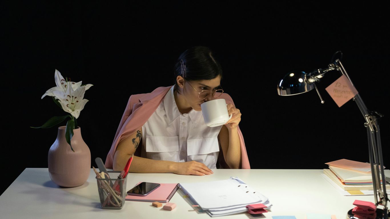 Person drinking coffee at a desk with a lily and lamp.