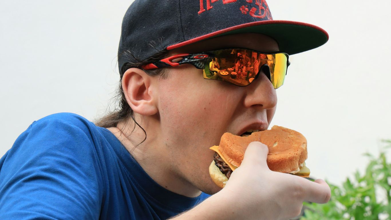 Man wearing sunglasses and a hat is eating a burger.