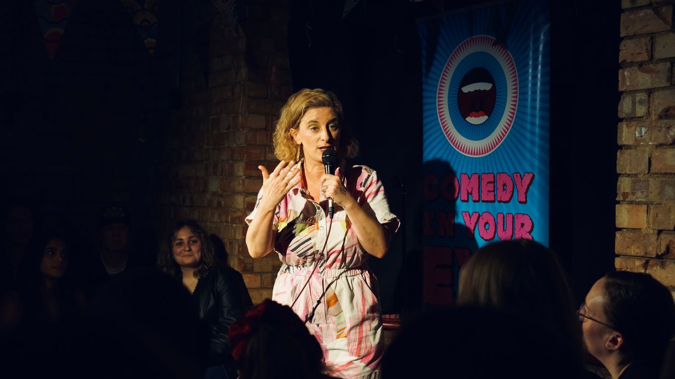 A person performs stand-up comedy in front of a banner.