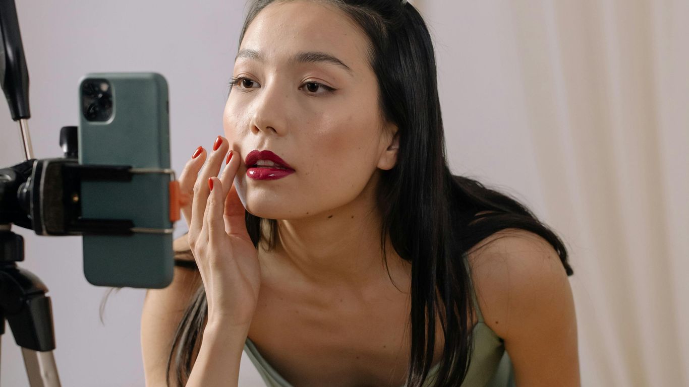 Person applies makeup while looking at smartphone on tripod stand.