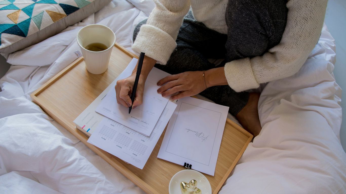 person in white sweater writing on white paper beside white ceramic mug on brown wooden table