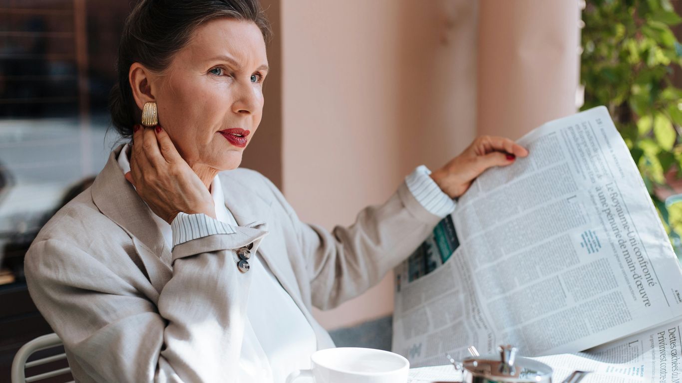 Person sitting at table, reading newspaper, drinking coffee, looking thoughtful.