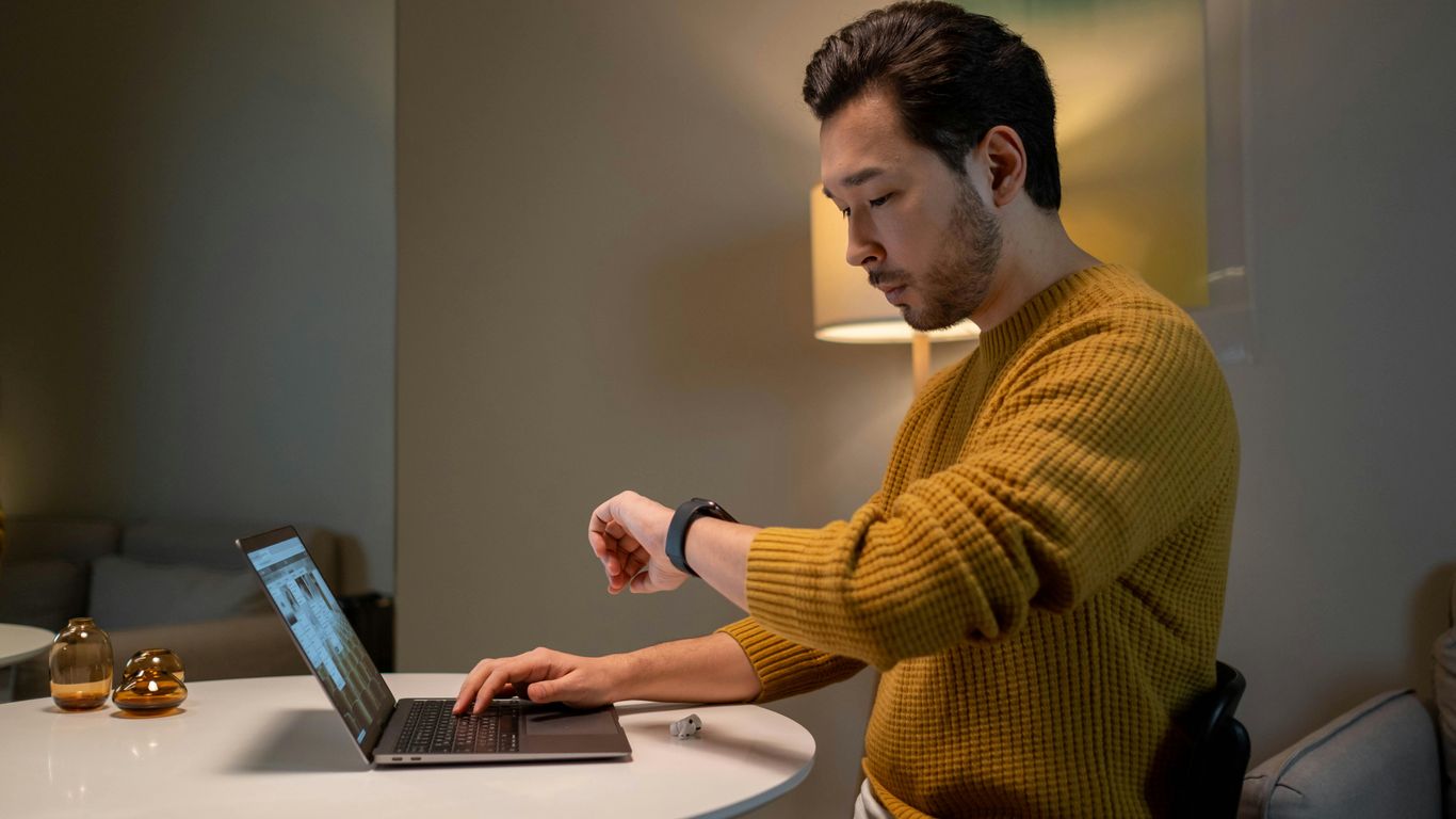 Person at desk using laptop, checking smartwatch, cozy indoor setting.