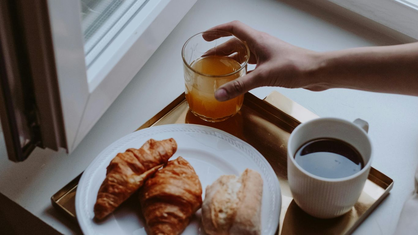 Breakfast tray with croissants, coffee, juice, and bread by window.