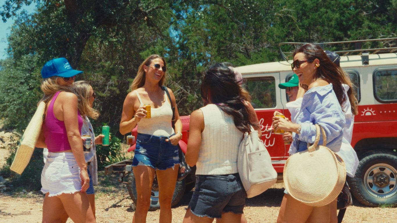 Group of people with drinks near a red vehicle in a wooded area.