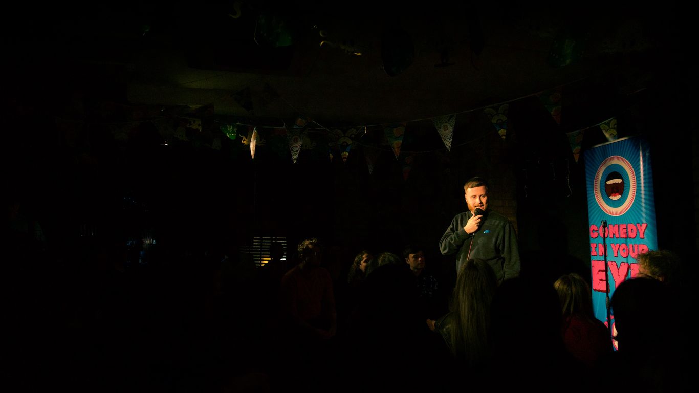 A comedian performs on stage with a banner that says "Comedy in Your Eye."