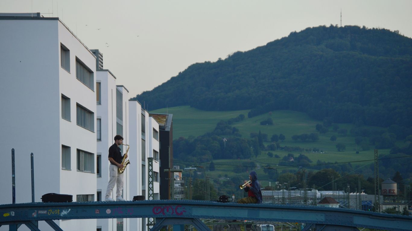 Man plays saxophone on bridge with buildings and mountain.
