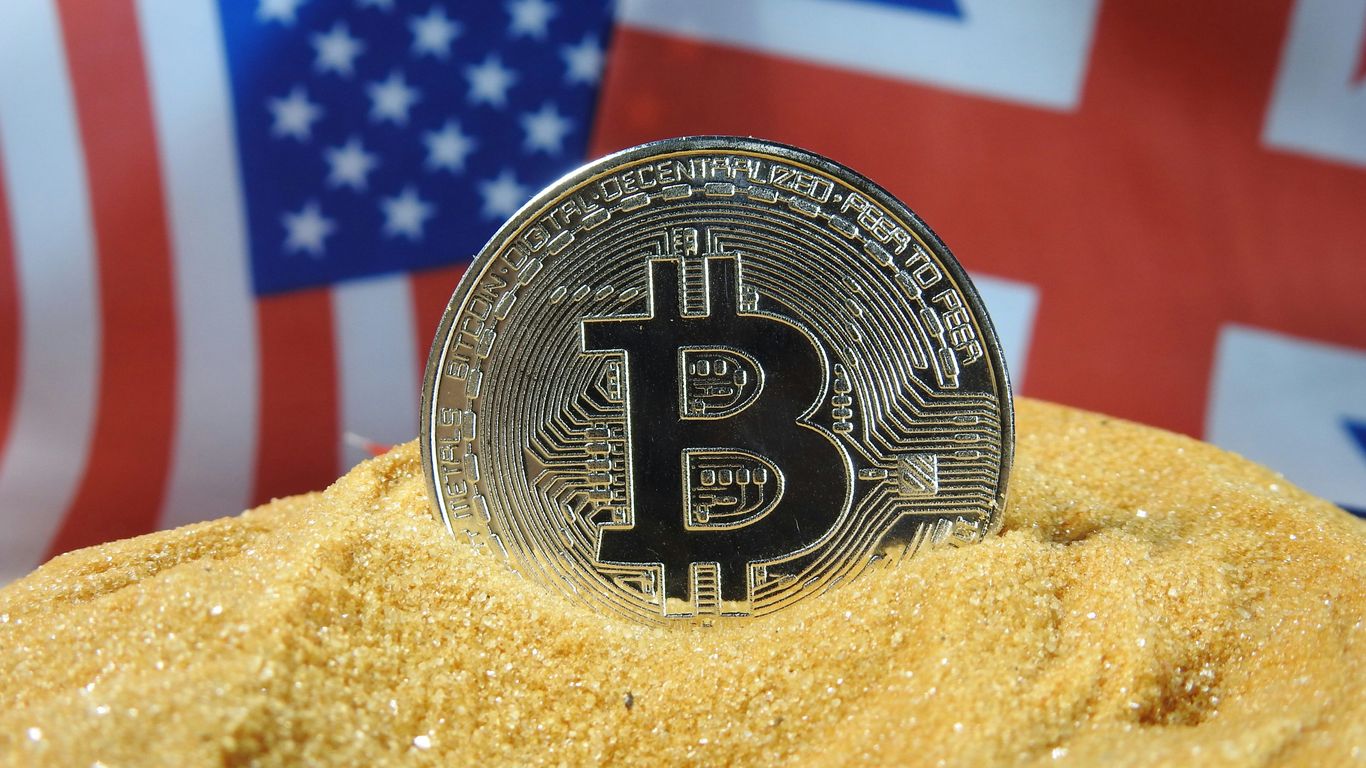 Bitcoin coin resting in sand with flags in background.