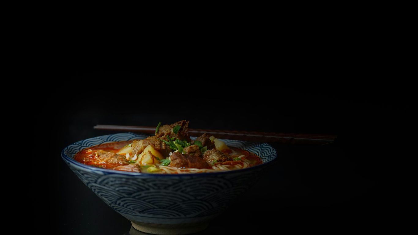 Bowl of spicy noodle soup with chopsticks, dark background.