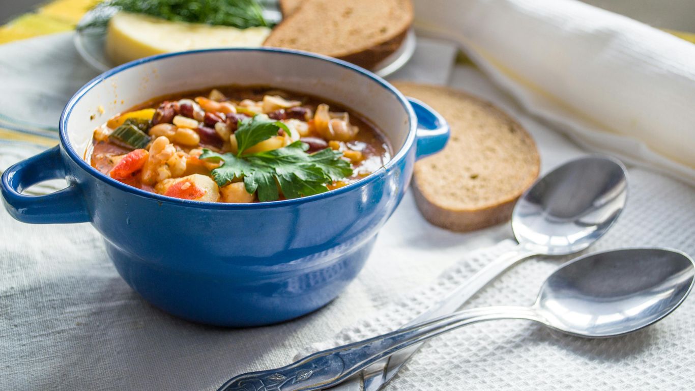 Hearty vegetable soup in blue bowl with bread and spoons.