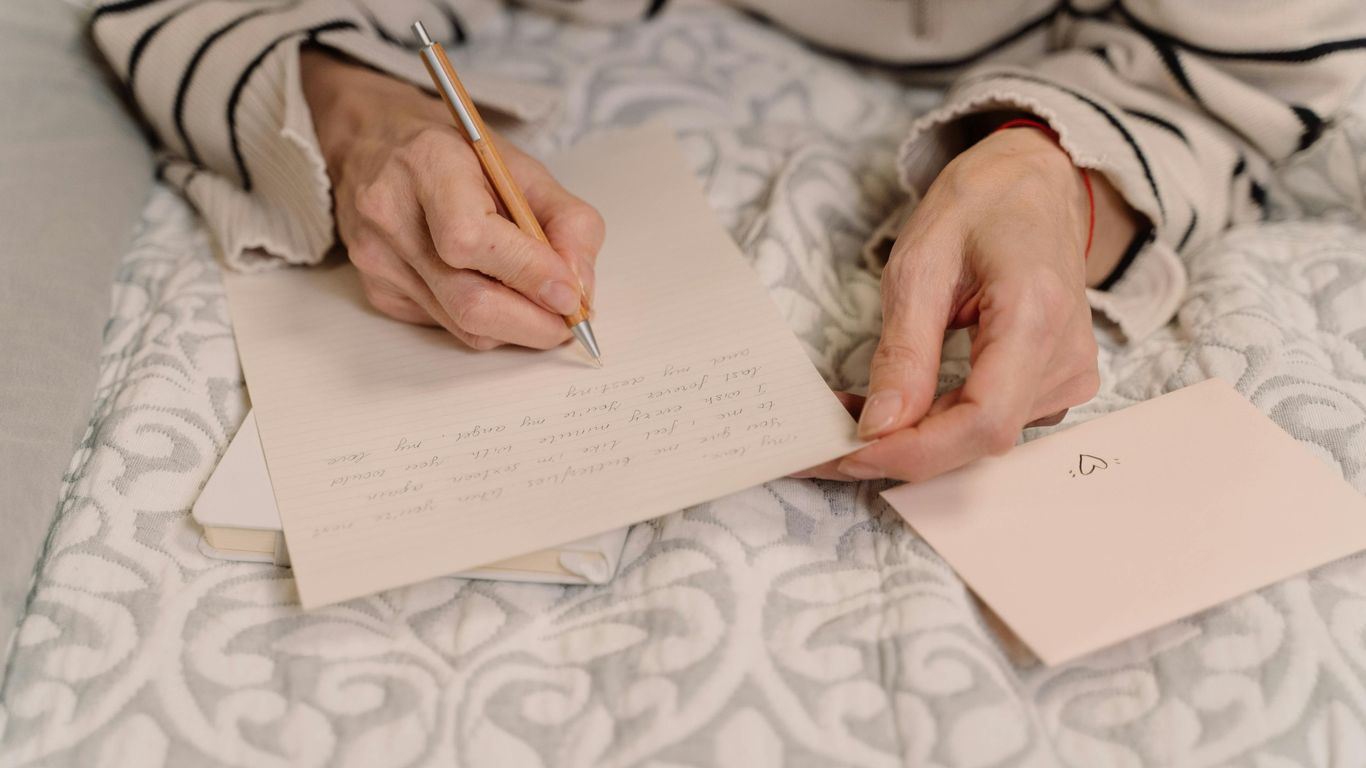 Person writing a letter on bed, envelope with heart nearby.