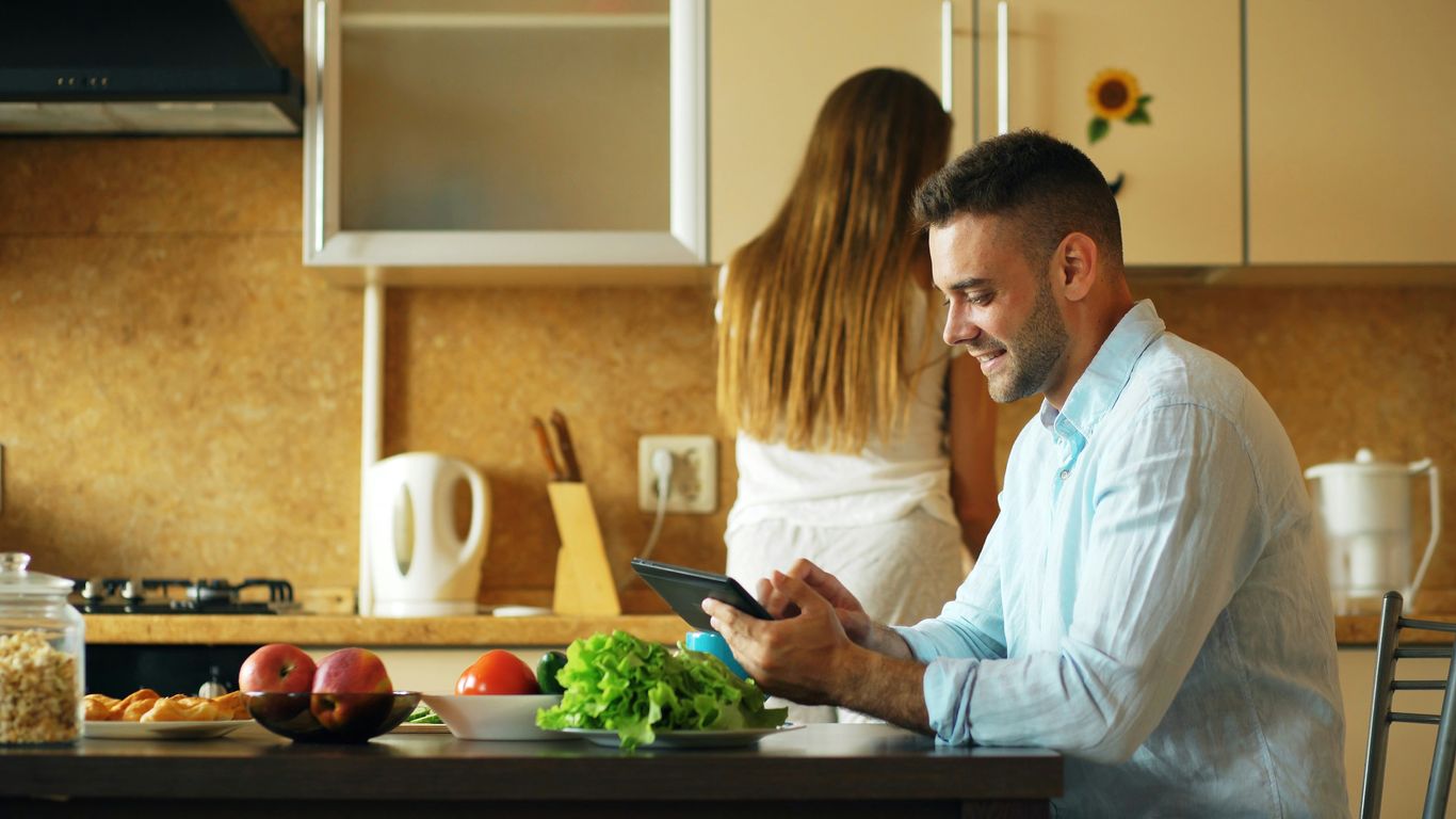 Man using tablet in kitchen while woman stands behind him.