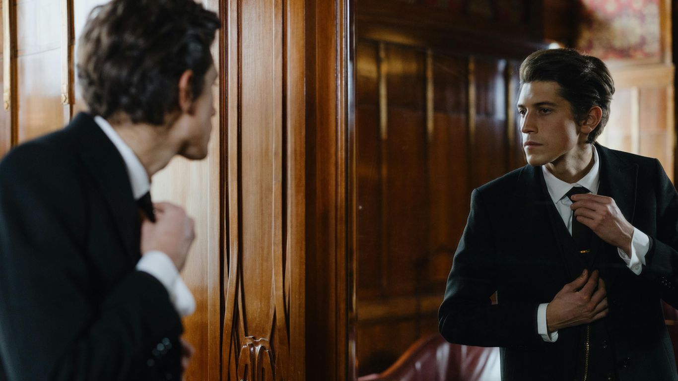Man in a suit adjusts his tie in a mirror.