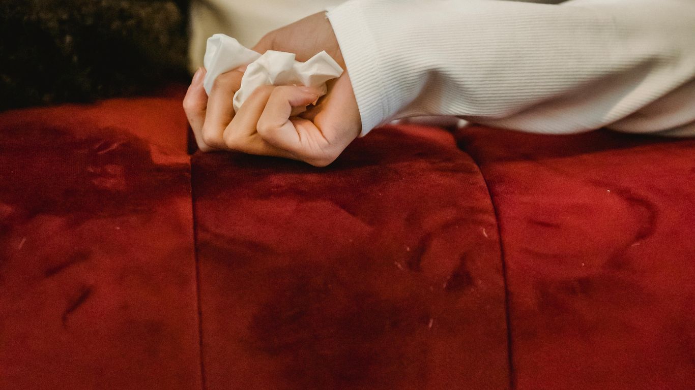 Person lying on red couch, holding tissue, wearing white shirt.