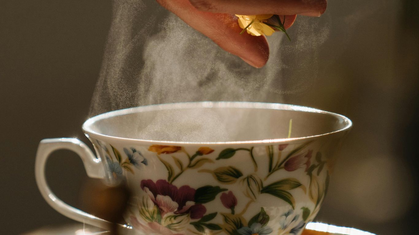 Hand dropping flower into steaming floral teacup, warm sunlight.