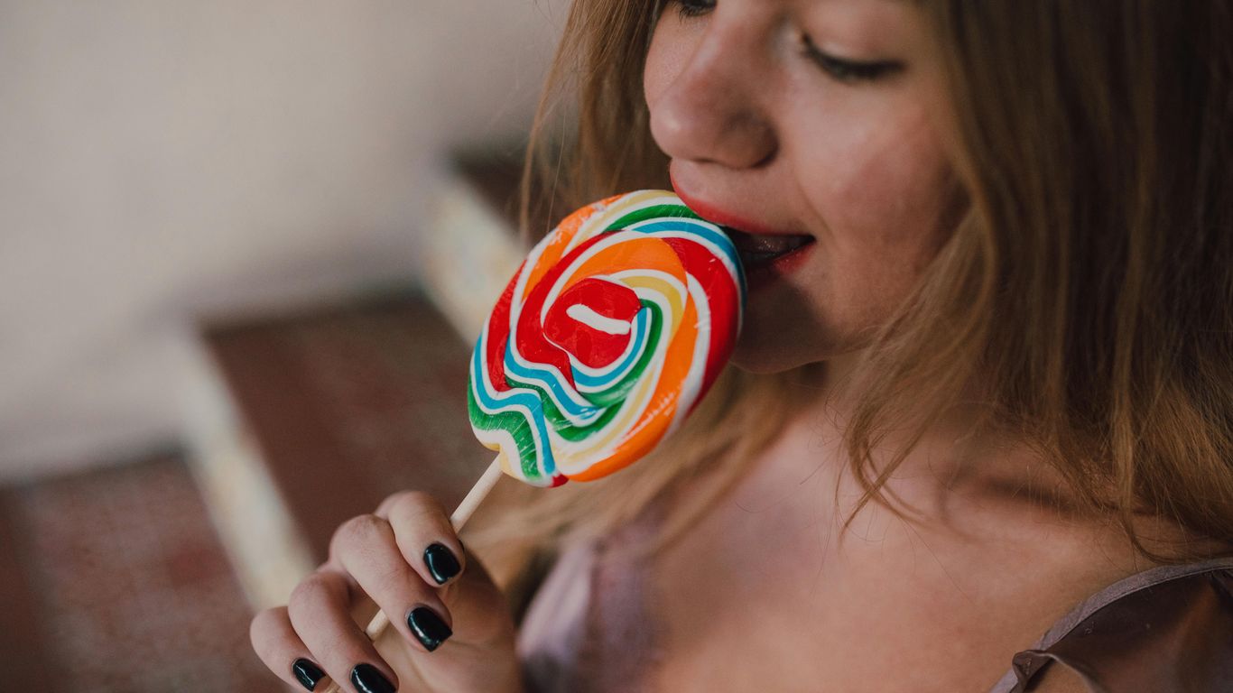 Person holding and licking a large colorful swirled lollipop.