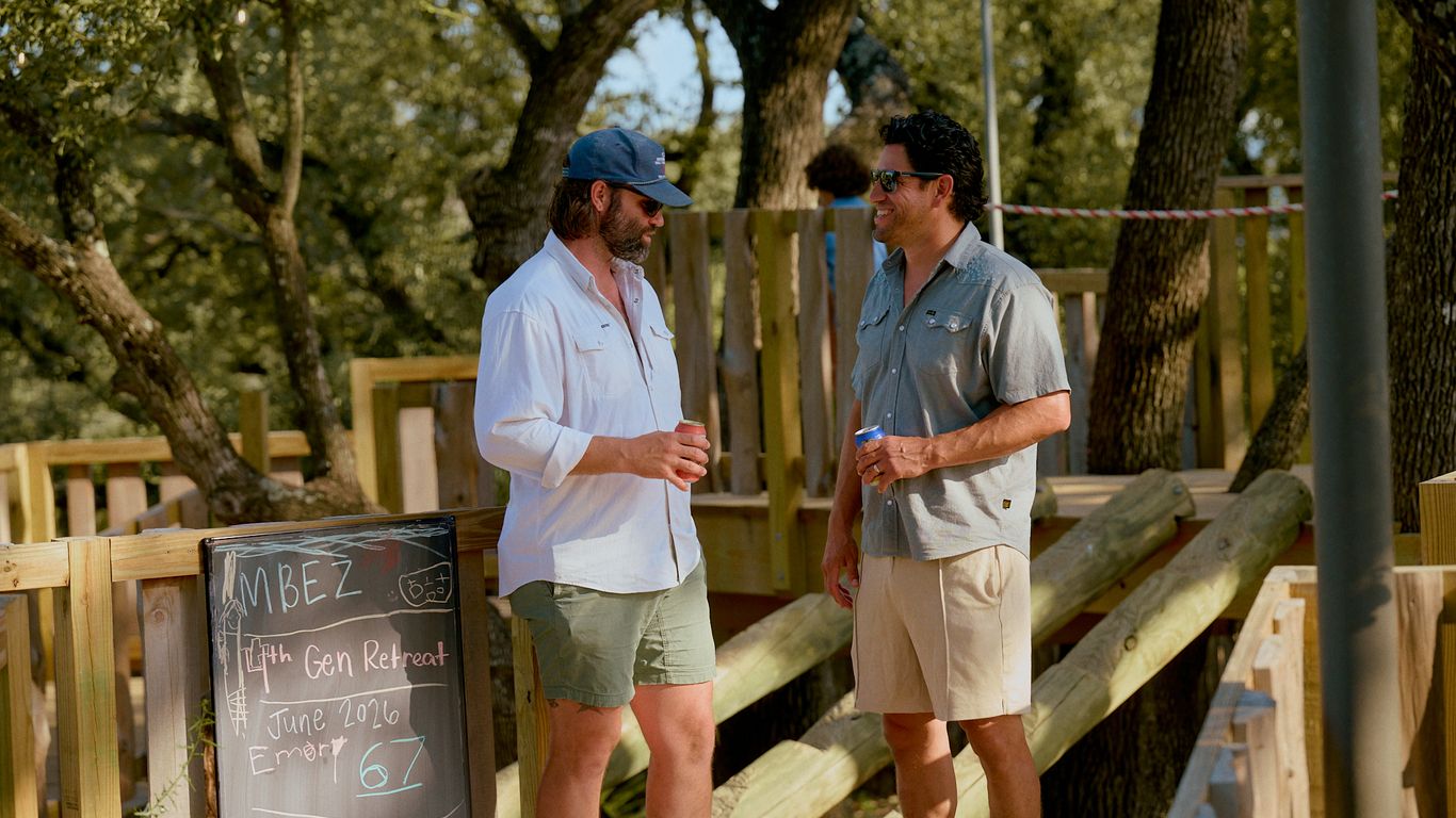 Two men talking outdoors near a wooden structure and trees.