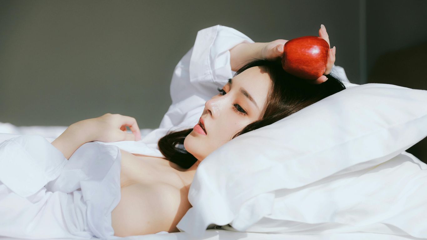 Person lying on bed, holding a red apple, white sheets.