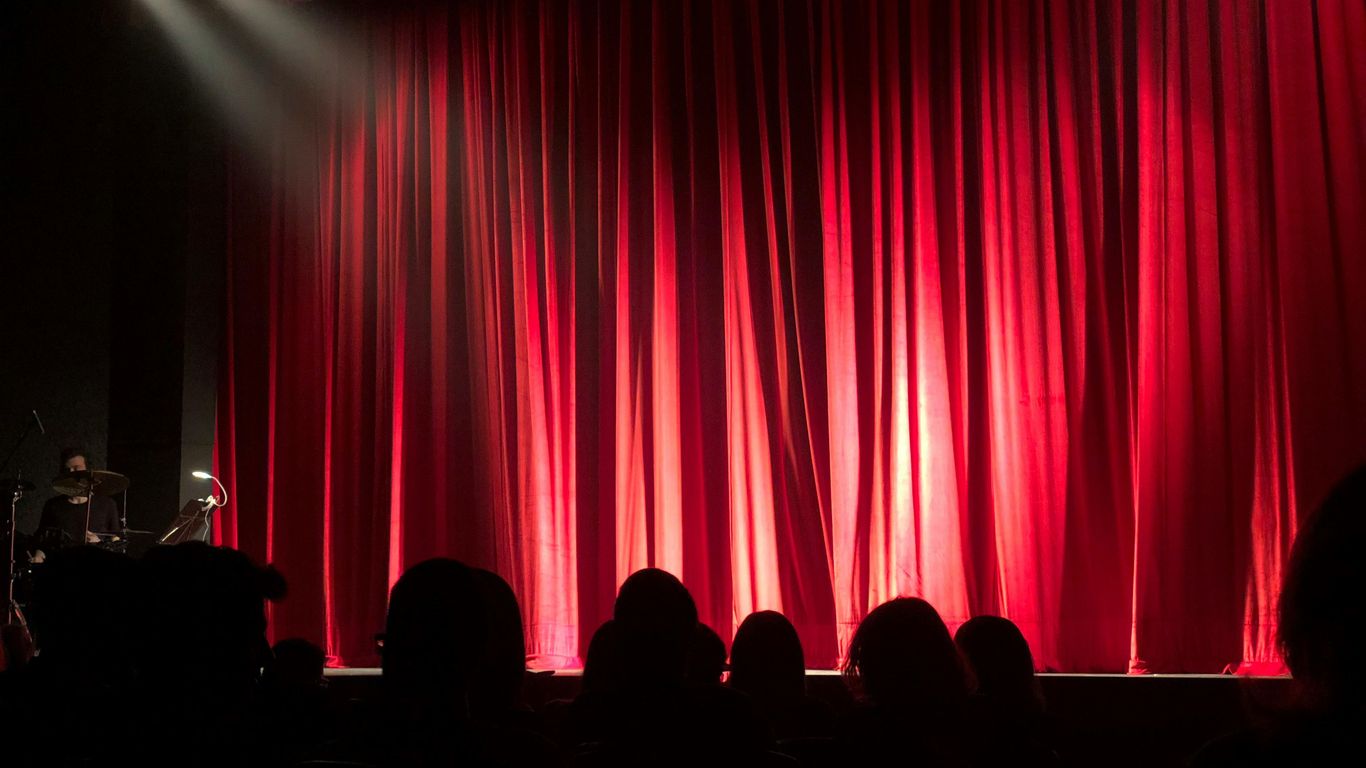 Theater stage with red curtains, audience in shadows waiting.