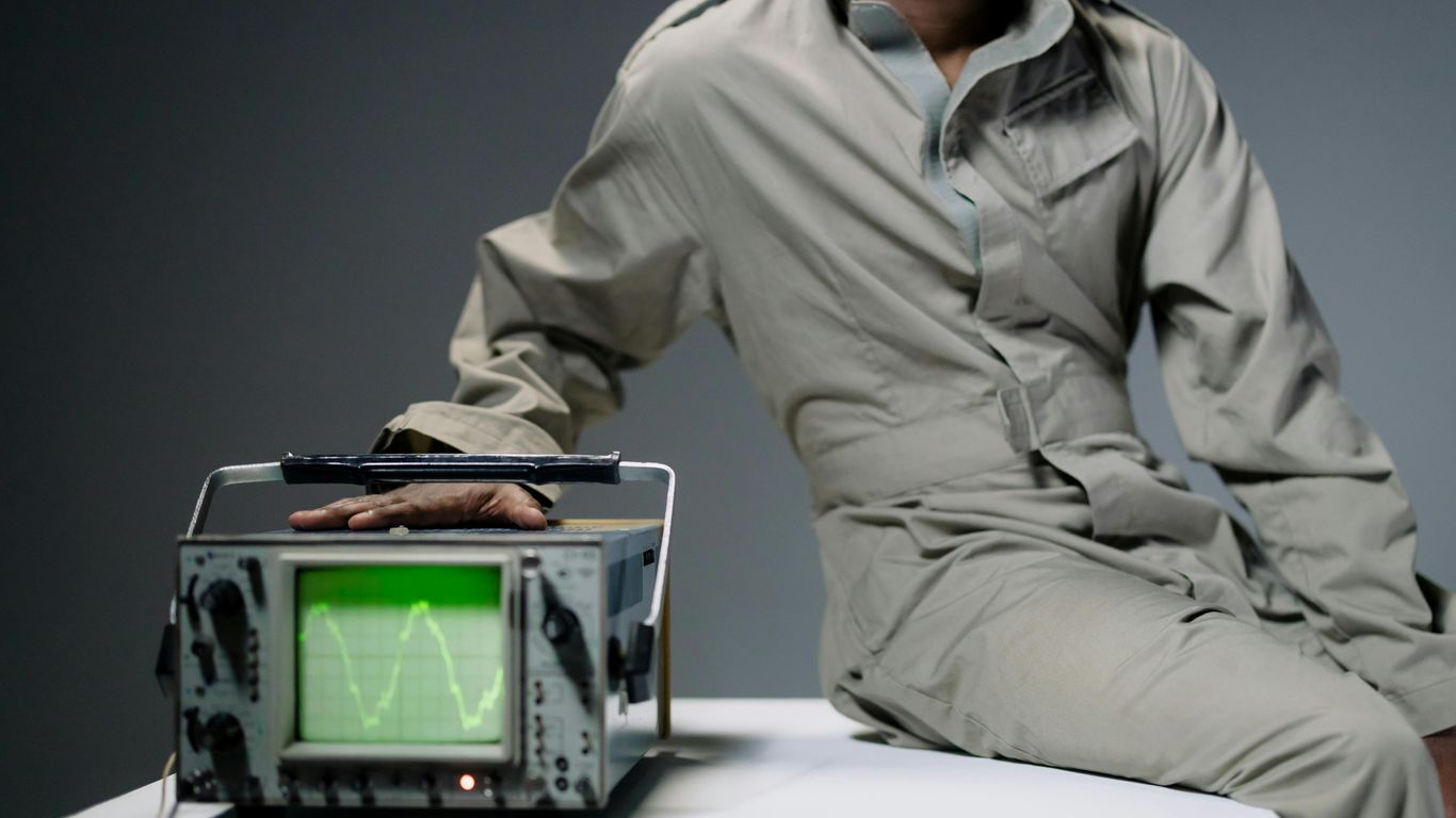 Person in jumpsuit with hand on oscilloscope displaying waveform.