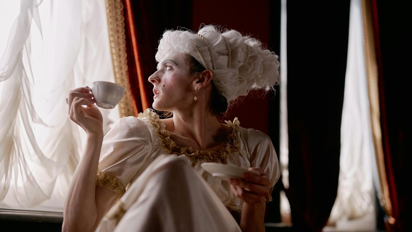 Person in historical costume drinking tea by elegant window curtains.